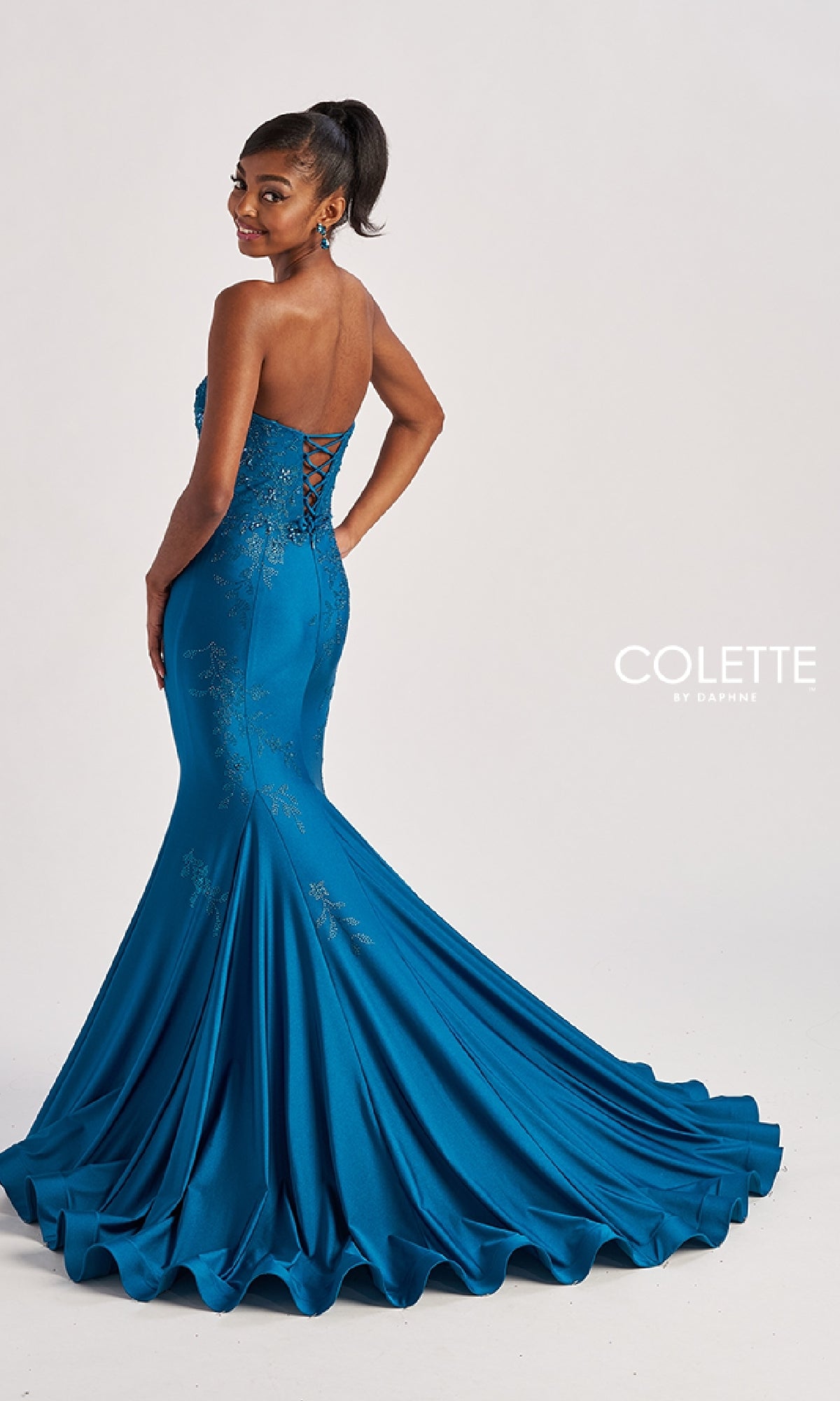A woman poses in the Colette CL8445 long prom dress, featuring blue sequins, a mermaid silhouette, sweetheart neckline, lace-up back, flared train, and subtle glittery patterns as she looks over her shoulder.